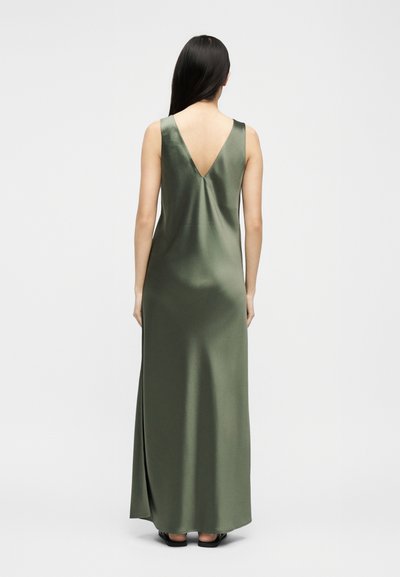 Γυναίκα με μακριά σκούρα μαλλιά, που φορά ένα αμάνικο, σατέν μακρύ φόρεμα σε ελιά πράσινο χρώμα με V-πλάτη, στέκεται μπροστά σε απλό ανοιχτόχρωμο φόντο.