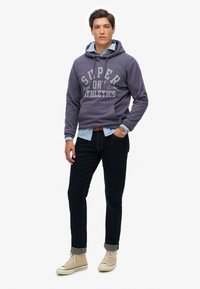 Sudadera con capucha gris con el texto "SUPER DRY ATHLETICS", camiseta azul debajo, pantalones oscuros y zapatillas altas color beige claro.