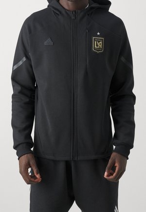 Sweat à capuche noir zippé avec le blason du Los Angeles Football Club et le logo Adidas, porté par une personne les mains détendues le long du corps.