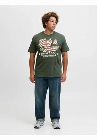 Camiseta de algodón verde con estampado blanco y rojo que dice "Jack & Jones" y "Denim Goods". Combinada con vaqueros de mezclilla azules y zapatillas deportivas grisáceas.