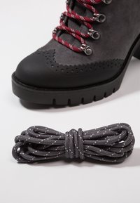 Botte de randonnée en suède gris avec lacets rouges et blancs, œillets en métal, et une bobine séparée de lacets gris foncé avec des taches blanches sur fond blanc.