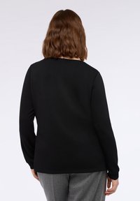 Eine Person mit schulterlangem braunem Haar, die ein langärmliges schwarzes Shirt und graue Hosen trägt und mit dem Rücken zu einer weißen Hintergrund steht.