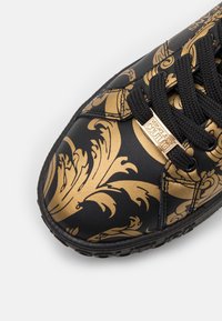 Baskets noires avec des motifs floraux baroques dorés, lacets noirs et une étiquette en métal doré indiquant "Versace Jeans Couture" à l'avant.