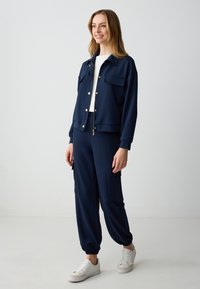 Completo felpa navy in tessuto morbido. La giacca ha dettagli con bottoni e tasche a toppa. I joggers presentano polsini elastici e tasche laterali.