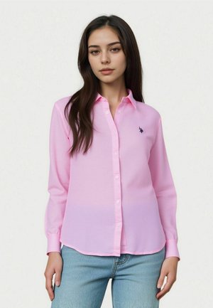 Donna con capelli lunghi e scuri che indossa una camicia rosa chiaro con bottoni e logo e jeans azzurri, in piedi contro uno sfondo uniforme.