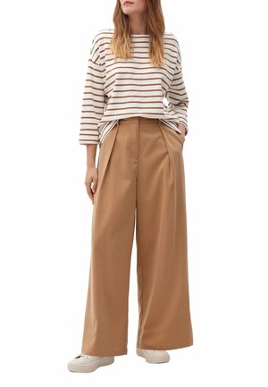 Mujer vestida con una camisa de manga larga a rayas blancas y beige, pantalones anchos de tiro alto color beige y zapatillas blancas, de pie con las manos en los bolsillos.