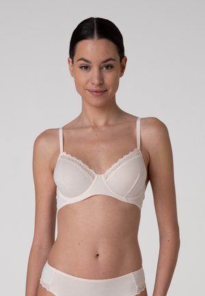 Donna con capelli scuri, indossa un reggiseno beige chiaro e biancheria intima coordinata, in piedi davanti a uno sfondo grigio chiaro uniforme.