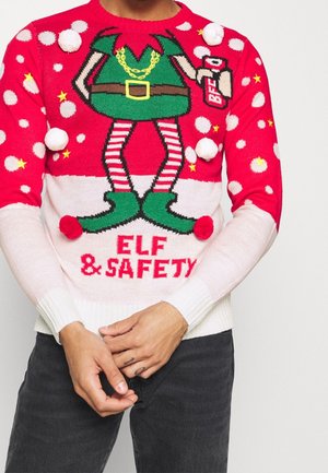 Rød og hvid julesweater med nisseben, pomponer, stjerner og teksten "Elf & Safety", båret af en person med hænderne foldet foran.