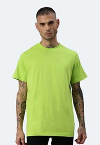 T-shirt in cotone verde neon con maniche corte, colletto rotondo e vestibilità rilassata. Presenta tatuaggi visibili sulle braccia.