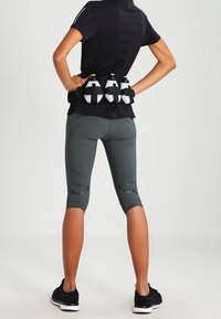 Czarna koszulka sportowa i szare legginsy z siateczkowymi akcentami. Trzy białe butelki na wodę przymocowane czarnymi paskami na plecach. Czarne sneakersy.