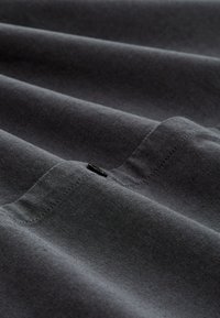 Schwarzes Hemd aus Stoff mit glatter Textur, sichtbaren Nähten und einem kleinen schwarzen Knopfdetail in der Nähe der Mitte.