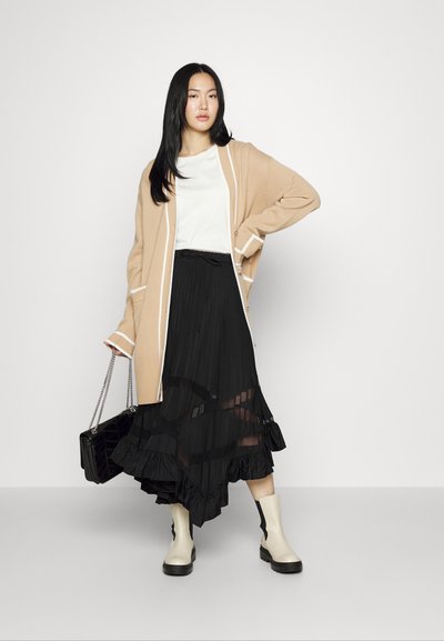 Beige gebreide cardigan met witte accenten, gelaagd over een zwarte plissérok met een rimpelige zoom, gecombineerd met crèmekleurige enkellaarsjes en een zwarte handtas.