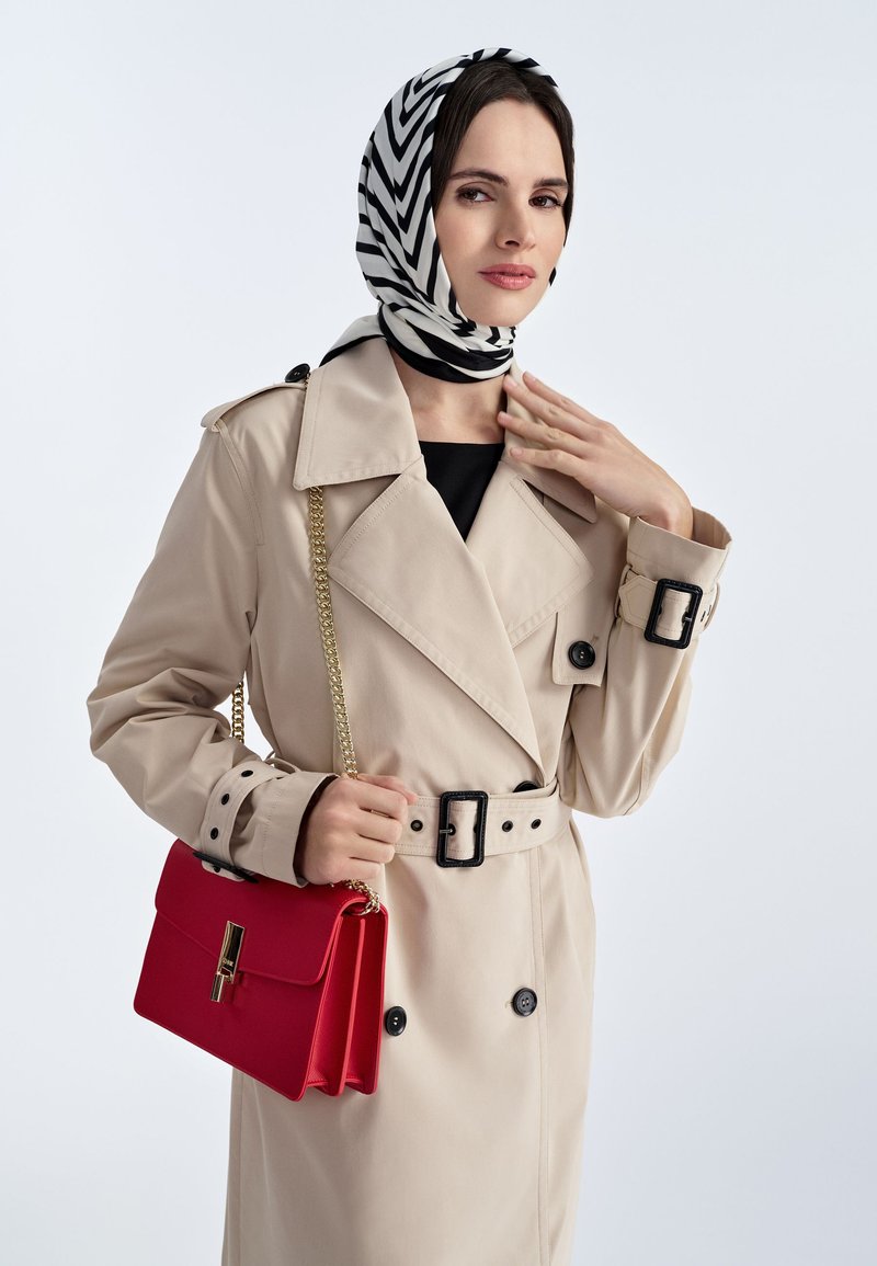Beżowy płaszcz trench z czarnymi guzikami i paskiem w talii; czerwona torebka z złotym łańcuszkiem; czarno-biały szalik w wzory.