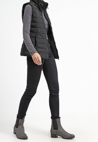 Černá prošívaná vesta přes šedé dlouhé tričko, spárovaná s černými slim džíny a šedými kotníkovými botami se bočními elastickými panely.