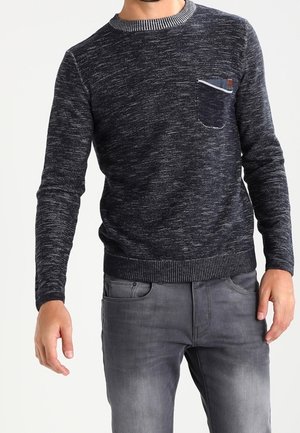 Ciemnoszary melanżowy sweter z dzianiny z prążkowanym kołnierzem, mankietami i obrębem, z kieszenią na piersi z małą skórzaną naszywką, noszony z wyblakłymi szarymi dżinsami.