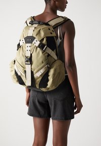 Grüner und schwarzer Rucksack aus strapazierfähigem Stoff, mit verstellbaren Trägern, Netzseitentaschen und einem modernen Design mit Schnallen.
