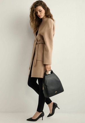 Kobieta w beżowym płaszczu z paskiem, czarnych spodniach i czarnych szpilkach trzyma czarną torebkę, stojąc na jasnym, jednolitym tle.