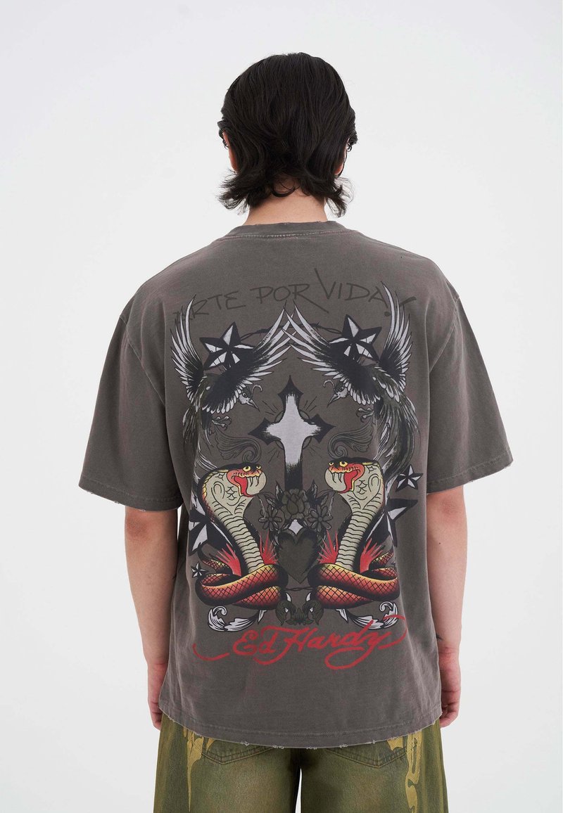 Šedé oversized tričko s barevným grafikou dvou hadů, křídel a hvězd na zádech. Krátké rukávy a kulatý výstřih.