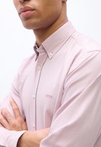 Άνδρας με σταυρωμένα τα χέρια, φοράει ανοιχτό ροζ πουκάμισο με κουμπιά και γιακά, με κεντημένο το λογότυπο "BOSS" στο στήθος.