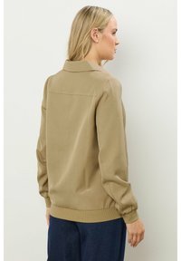 Giacca marrone chiaro con colletto, polsini elasticizzati e superficie liscia, indossata con pantaloni blu navy. Design semplice e pulito.