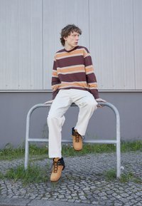Brun og oransje stripet langermet skjorte, hvite bukser og brune snørestøvler. Sitter på et metallskinner, uformell utendørs setting.