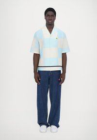 Camisa de manga corta abotonada en un patrón de cuadros azul claro y crema con un icono de sonrisa en el pecho, combinada con unos vaqueros de denim azul holgados.