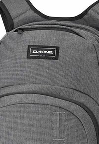 Grå ryggsäck med en texturerad tyg, utrustad med en framträdande svart etikett med "DAKINE." Inkluderar böjd dragkedja och vadderade axelremmar.