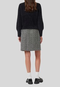 Frau, die einen schwarzen, flauschigen Pullover, einen knielangen Rock mit schwarz-weißen Mustern, weiße Socken und schwarze klobige Schuhe trägt und mit dem Rücken steht.