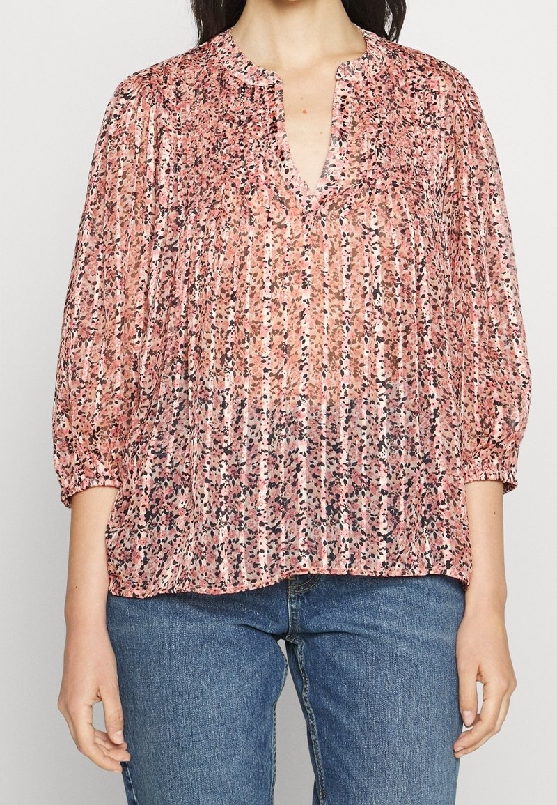 Kobieta ubrana w różową bluzkę w wzory z dekoltem w serek i rękawami o długości do łokcia z bufkami, zestawioną z niebieskimi dżinsami.