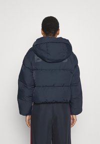 Doudoune bleu marine avec capuche, présentant une silhouette carrée, un design matelassé et un grand logo au dos.