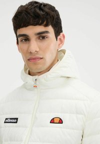 Chaqueta acolchada blanca con capucha, que cuenta con puños elásticos, un cierre frontal y el logo de Ellesse en el pecho. Detalle del logo en contraste en la capucha.