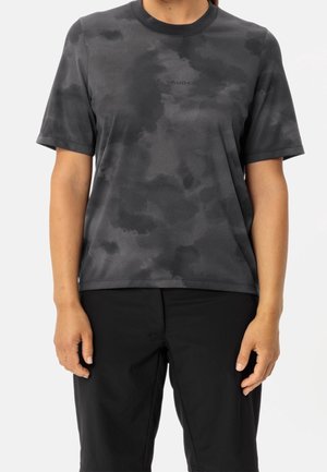 Persoon draagt een zwart met donkergrijs tie-dye patroon t-shirt met korte mouwen en een zwarte broek, staand met ontspannen armen.