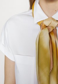 Camisa blanca de manga corta con bolsillo, que presenta una bufanda de satén atada de manera suelta en tonos de oro y marrón alrededor del cuello.