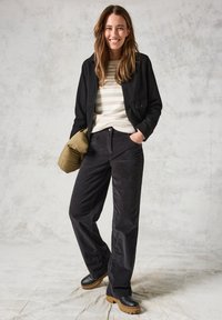 Pantalons en velours noir, pull à rayures crème, veste noire. Chaussures à plateforme marron. Le mannequin tient un sac doudoune vert olive. Fond neutre.