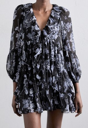 Czarna sukienka w kwiaty z falbankowym dekoltem w kształcie litery V, długimi bufkami i warstwową spódnicą, noszona przez stojącą osobę na jednolitym tle.