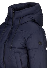 Marineblauwe gewatteerde jas met hoge kraag, ritssluiting aan de voorkant, trekkoorden en een patch op de mouw. De stof lijkt gewatteerd en zacht.