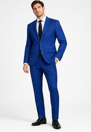Homme portant un costume bleu vif ajusté, chemise blanche, cravate noire, ceinture noire et chaussures habillées noires, debout avec une main dans la poche.
