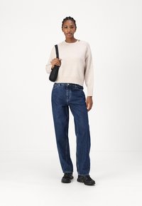 Beige strikket genser med rund hals og knappedetalj, kombinert med blå høytlivs straight-leg jeans og svarte joggesko.