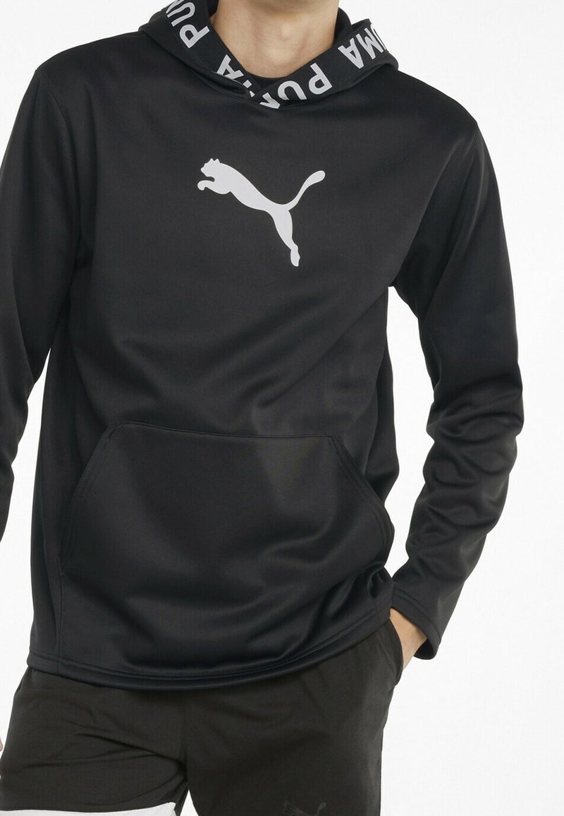 Homme portant un sweat à capuche noir Puma avec poche avant et logo sur la poitrine, associé à un short noir Puma.