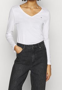 Witte langearmige t-shirt met een V-hals en een klein logo op de linkerkant van de borst, gecombineerd met donkergrijze high-waisted jeans.
