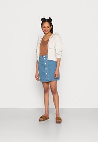 Jacket branco com capuz sobre uma blusa castanha, combinada com uma saia de ganga azul com botões na frente. Sandálias de cunha castanhas apresentam um detalhe em nó.