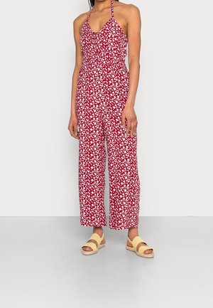 Kvinde iført ærmeløs rød jumpsuit med hvidt blomsterprint og gule åbne sandaler, stående mod hvid baggrund.
