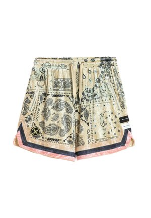 Persona rubia de pie con camisa grande beige con patrón de cachemira y pantalones cortos a juego, calcetines blancos y zapatos beige gruesos sobre fondo blanco.