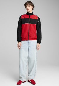 Giacca rossa e nera con zip, con dettagli bianchi, che presenta il logo Ferrari. Il modello indossa jeans azzurri chiari e sneakers rosse. Materiale liscio e sportivo.
