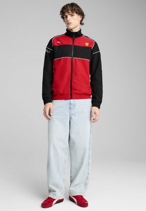 Giacca rossa e nera con zip, con dettagli bianchi, che presenta il logo Ferrari. Il modello indossa jeans azzurri chiari e sneakers rosse. Materiale liscio e sportivo.