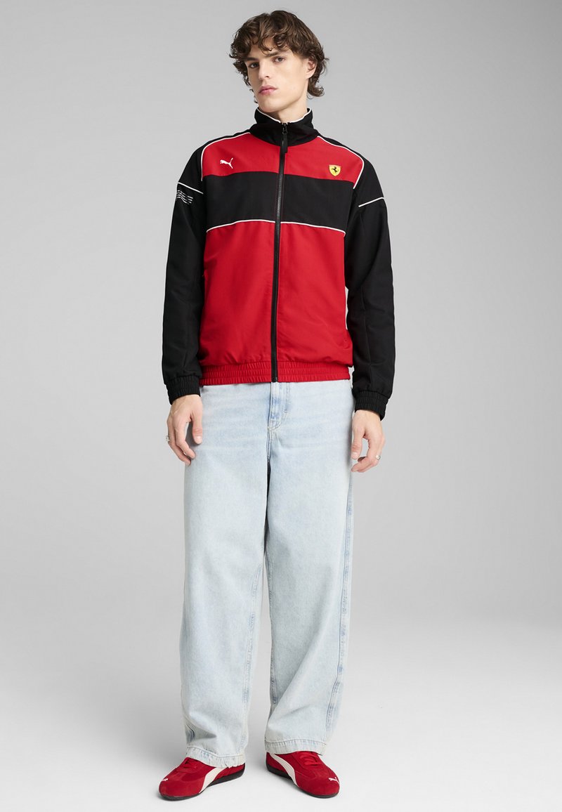 Giacca rossa e nera con zip, con dettagli bianchi, che presenta il logo Ferrari. Il modello indossa jeans azzurri chiari e sneakers rosse. Materiale liscio e sportivo.
