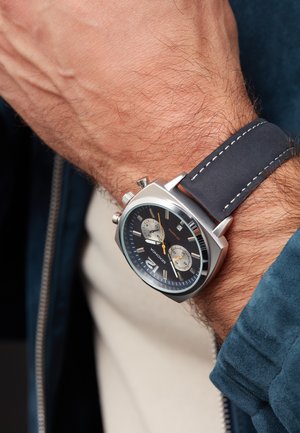 Chronograph watch with a silver stainless steel case, black dial, two sub-dials, and navy leather strap with white stitching.