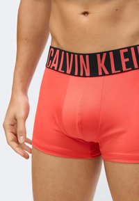 Mies torso, joka on pukeutunut kirkkaan punaisiin bokserihousuihin, joissa on musta vyötärönauha, jossa on suuri punainen "Calvin Klein" -logo.