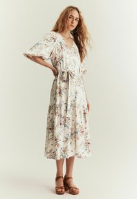 Blomstret midi-kjole med v-hals, korte puffærmer og bindebælte. Stoffet er let og har en hvid baggrund med pink og blå blomster.