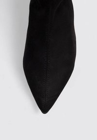 Zapato negro de ante con punta afilada y silueta estilizada, con costuras visibles. Textura suave, sin herrajes ni adornos.